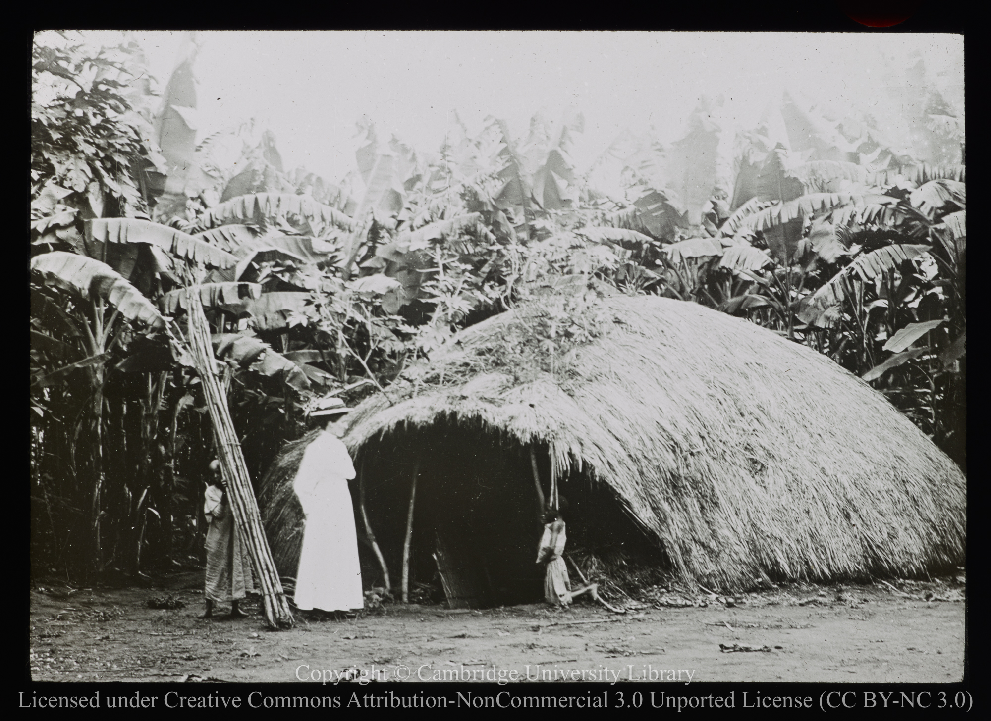 European woman standing outside grass hut, 1892 - 1914