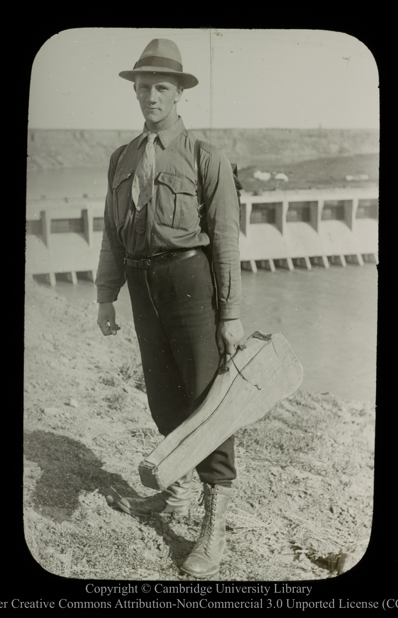 [Young man with gun(?) case, with dam in background], 1910 - 1930